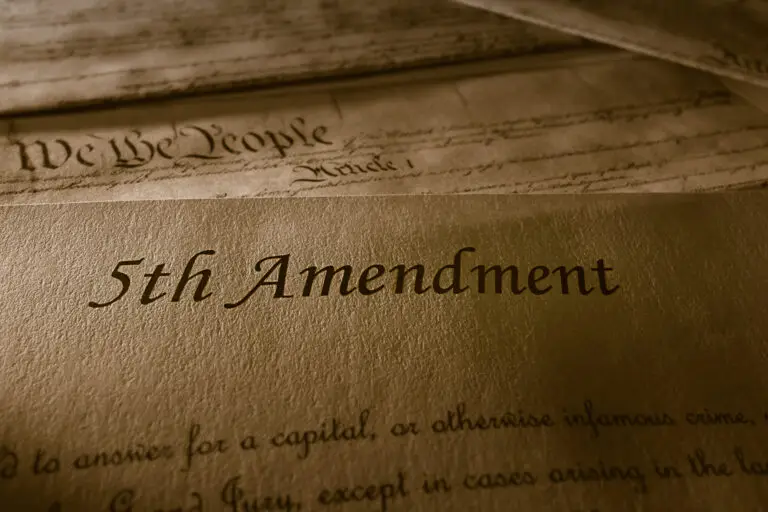 Close-up photo of a document from the bill of rights with "5th Amendment" prominently displayed, with the top edge of the United States Constitution showing "We the People" in the background.